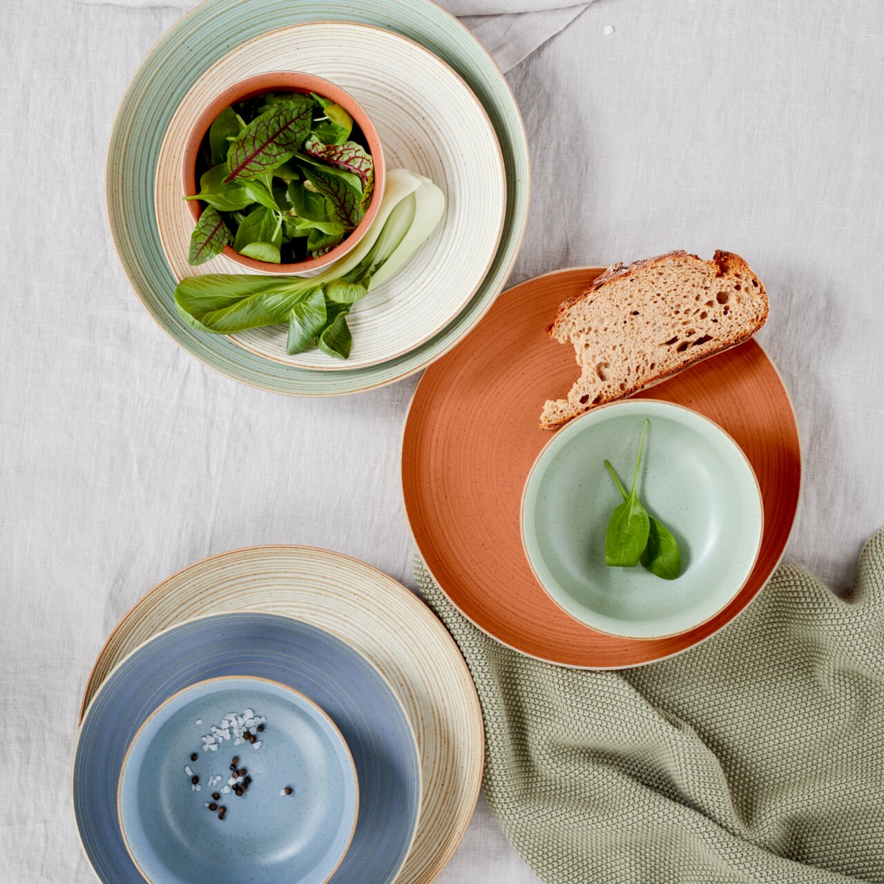 Thomas Ontbijtbord NATURE Vogelperspektive auf eine Auswahl von Frühstückstellern in verschiedenen Farben, darunter Grün, Blau und Terrakotta, mit frischen Zutaten wie Salat und Brot.