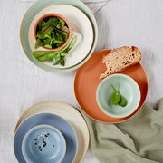 Vogelperspektive auf eine Auswahl von Frühstückstellern in verschiedenen Farben, darunter Grün, Blau und Terrakotta, mit frischen Zutaten wie Salat und Brot.