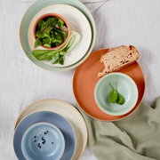 Vogelperspektive auf eine Auswahl von Frühstückstellern in verschiedenen Farben, darunter Grün, Blau und Terrakotta, mit frischen Zutaten wie Salat und Brot.