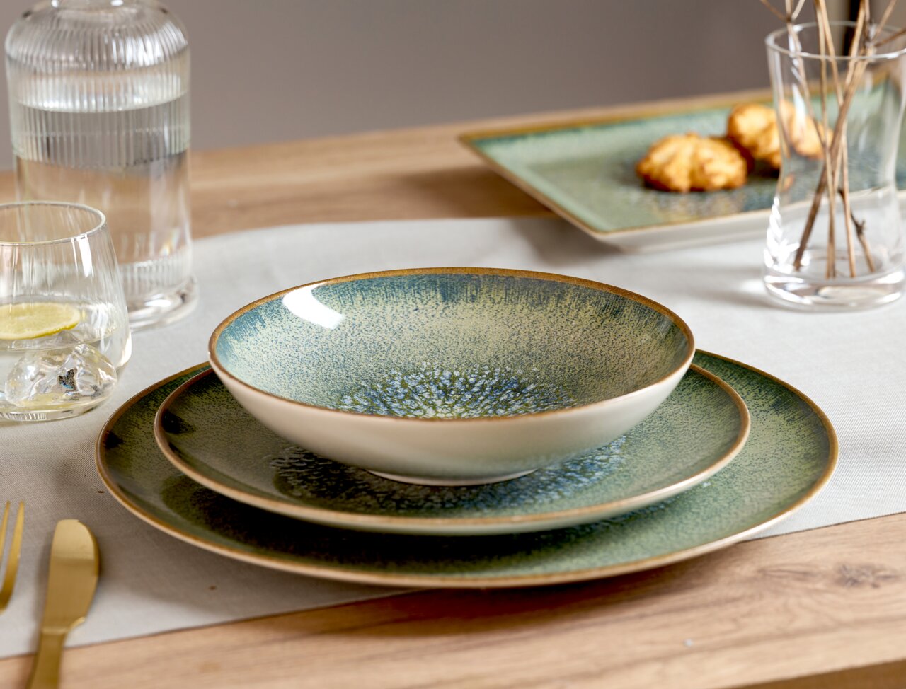 Een servies van keramiek in groenblauw met gespikkelde glazuur en een bruine rand, schuin van voren gefotografeerd: een diep bord/schaal op twee bijpassende borden op een gedekte houten tafel.