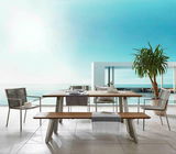 Moderner Gartentisch mit Holzplatte und Metallbeinen, umgeben von stilvollen Stühlen und einer Bank, auf einer Terrasse mit Meerblick, aus frontaler Perspektive.