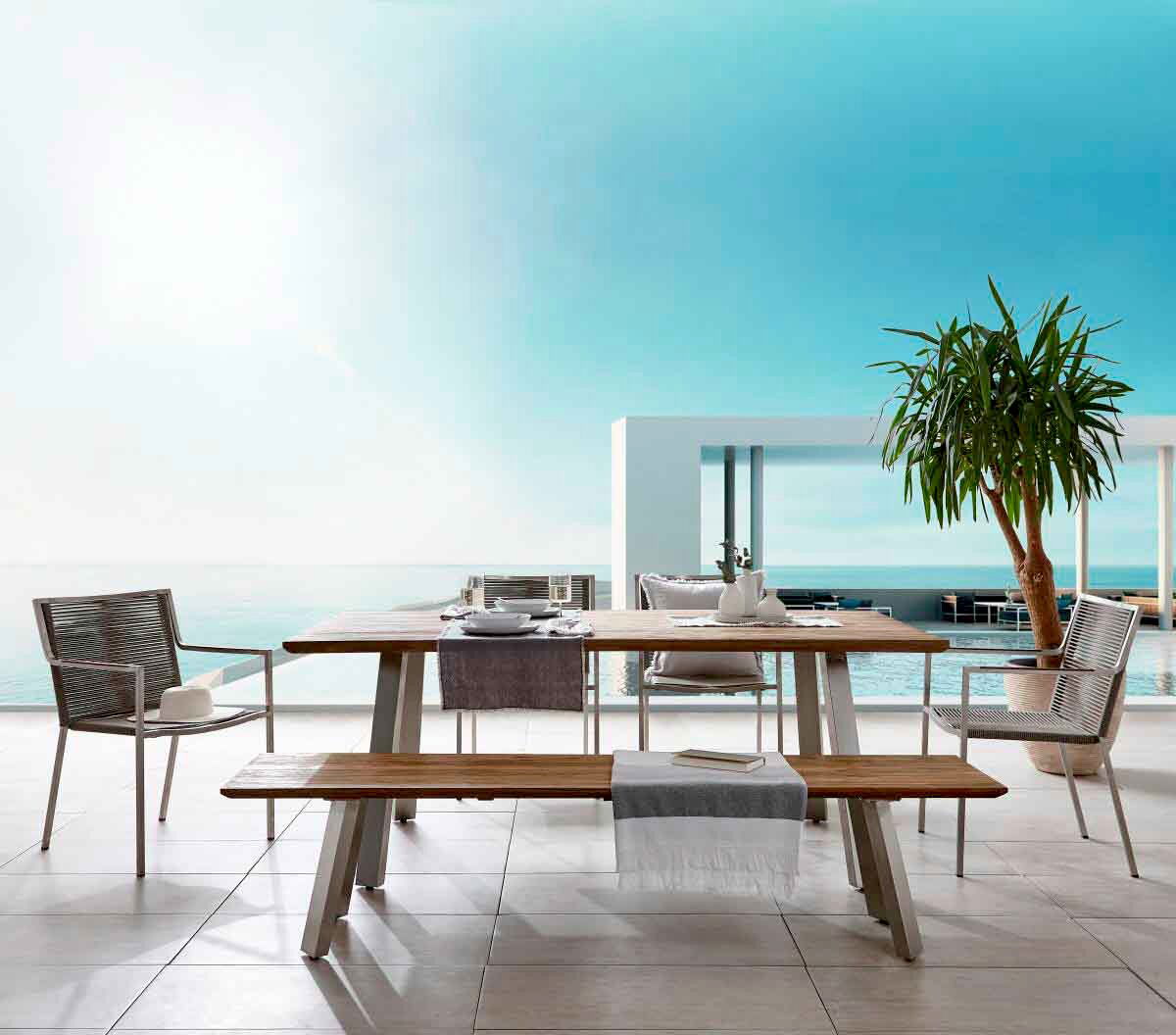 Amatio Tuintafel EPIRUS II Moderner Gartentisch mit Holzplatte und Metallbeinen, umgeben von stilvollen Stühlen und einer Bank, auf einer Terrasse mit Meerblick, aus frontaler Perspektive.