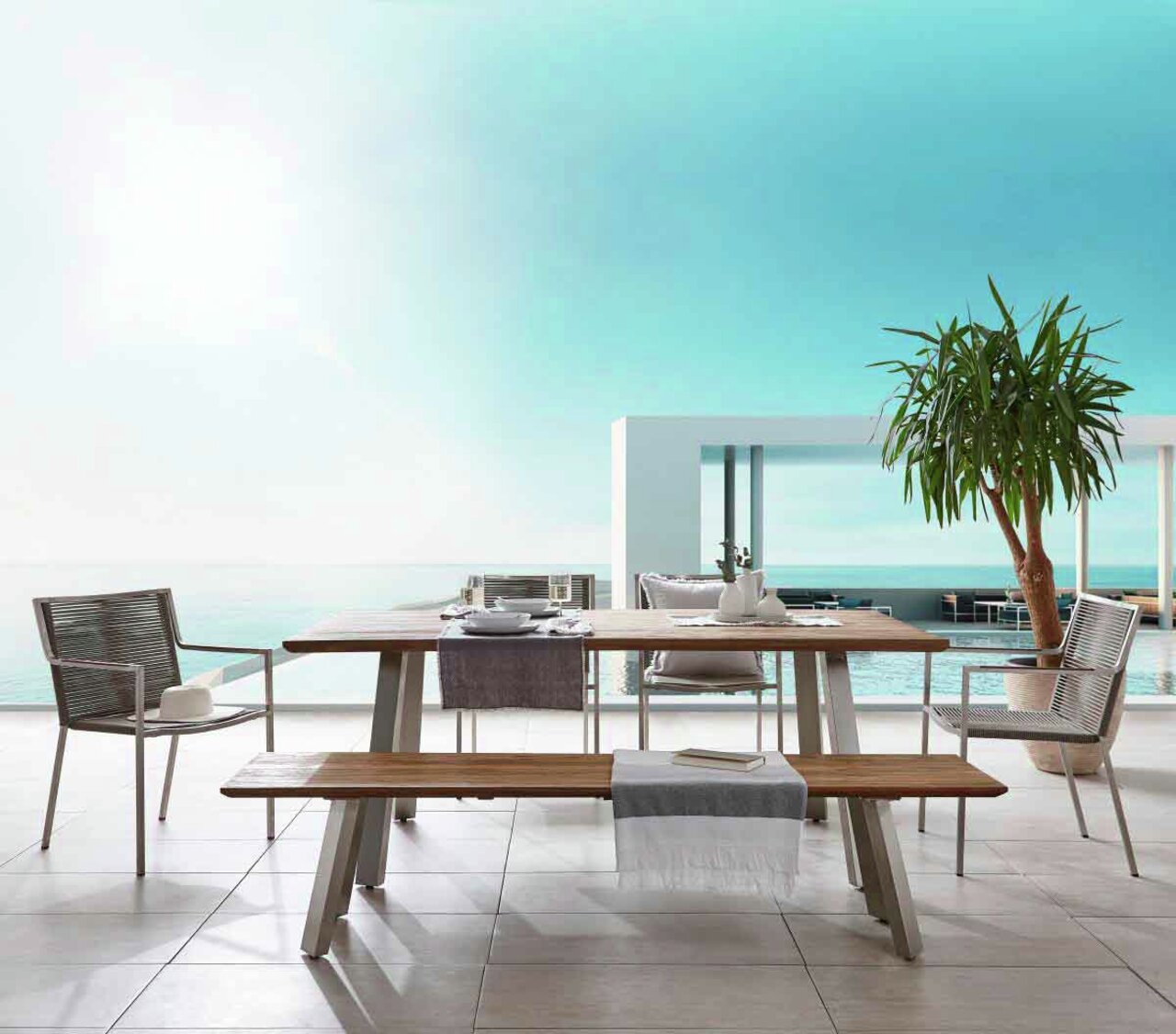 Amatio Gartentisch EPIRUS II Moderner Gartentisch mit Holzplatte und Metallbeinen, umgeben von stilvollen Stühlen und einer Bank, auf einer Terrasse mit Meerblick, aus frontaler Perspektive.