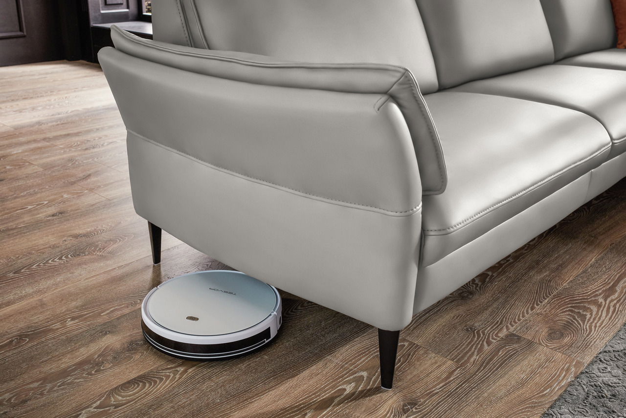 Graues Sofa mit einem Saugroboter, der darunter hervorschaut, auf einem Holzboden. Perspektive von der Seite.