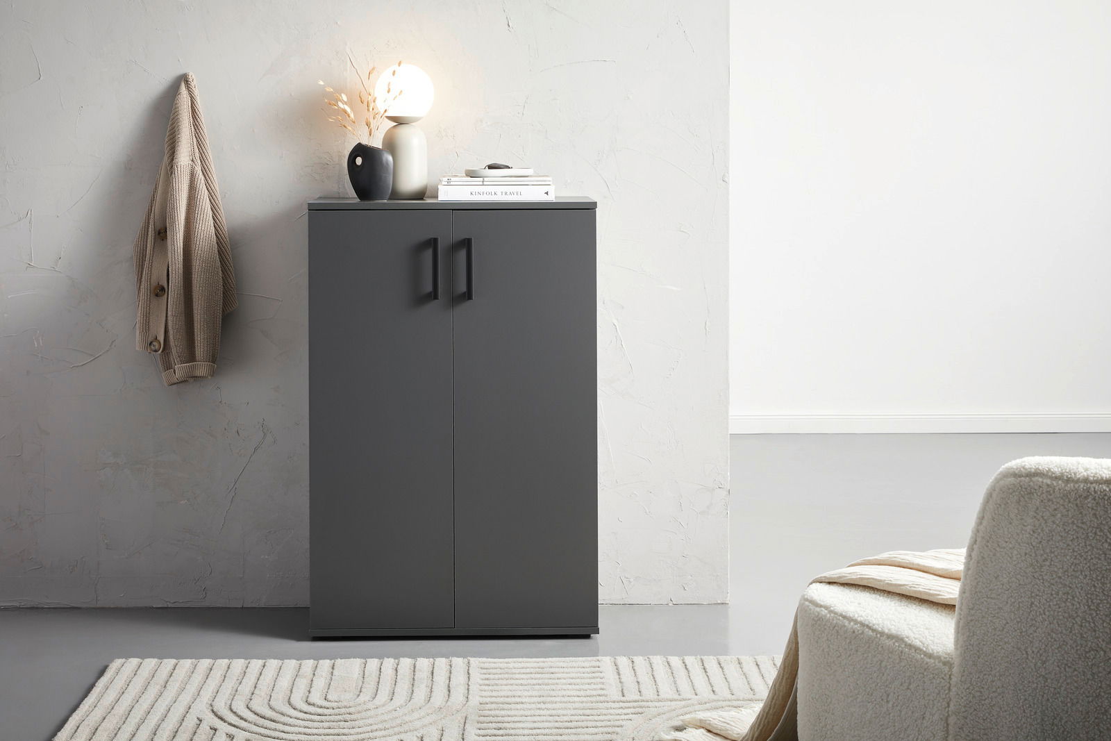 Moderner grauer Schuhschrank mit zwei Türen, frontal fotografiert, dekoriert mit einer Lampe und Vasen auf der Oberseite, neben einer beigen Jacke an der Wand.