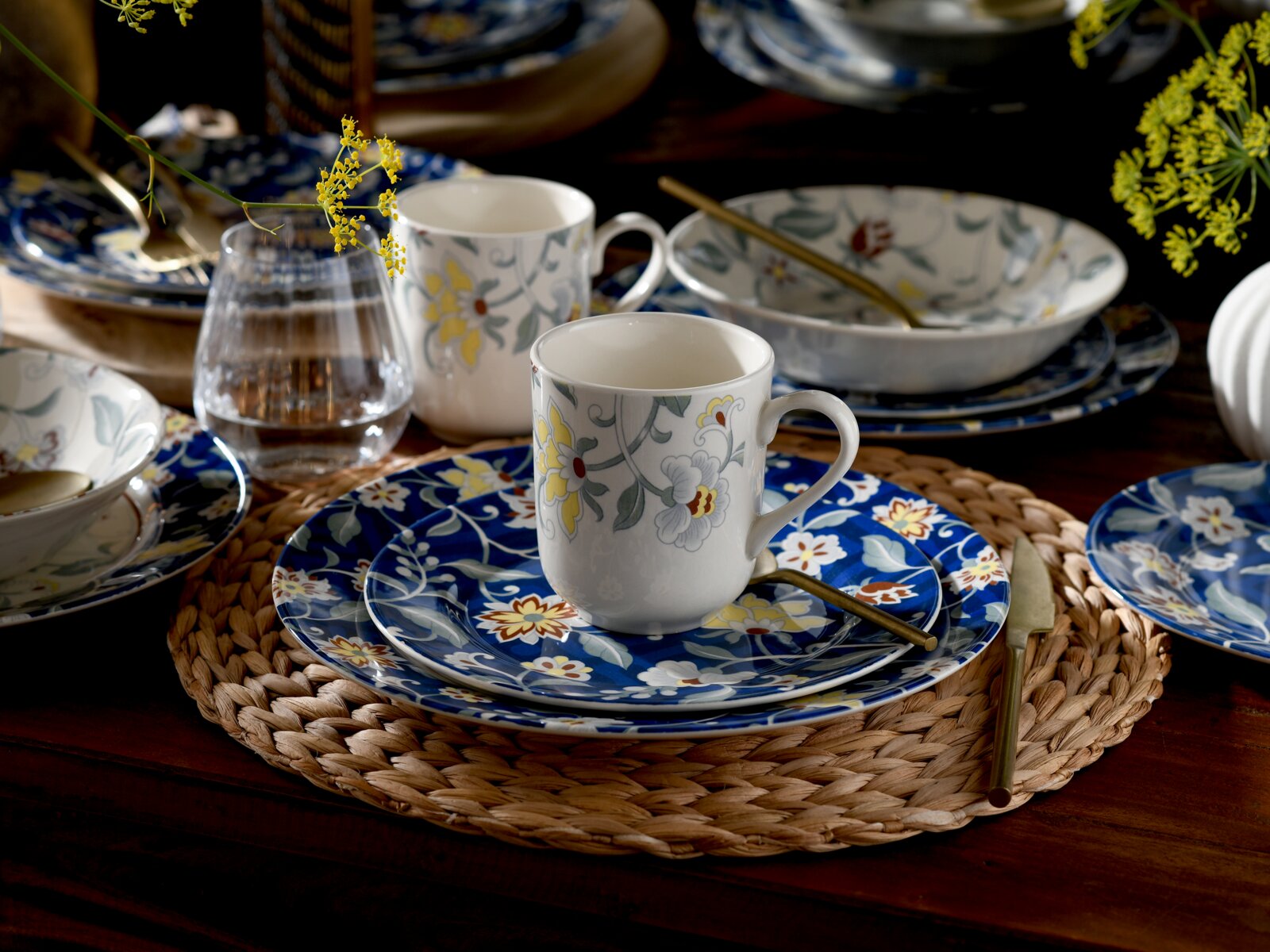 Gedeckter Esstisch mit blauem, floral gemustertem Keramikgeschirr und weißen Tassen mit Blumenmotiv, Glas und goldfarbenem Besteck auf geflochtenem Platzset, aufgenommen aus leicht erhöhter schräger Vorderansicht.