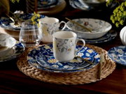 Gedeckter Esstisch mit blauem, floral gemustertem Keramikgeschirr und weißen Tassen mit Blumenmotiv, Glas und goldfarbenem Besteck auf geflochtenem Platzset, aufgenommen aus leicht erhöhter schräger Vorderansicht.
