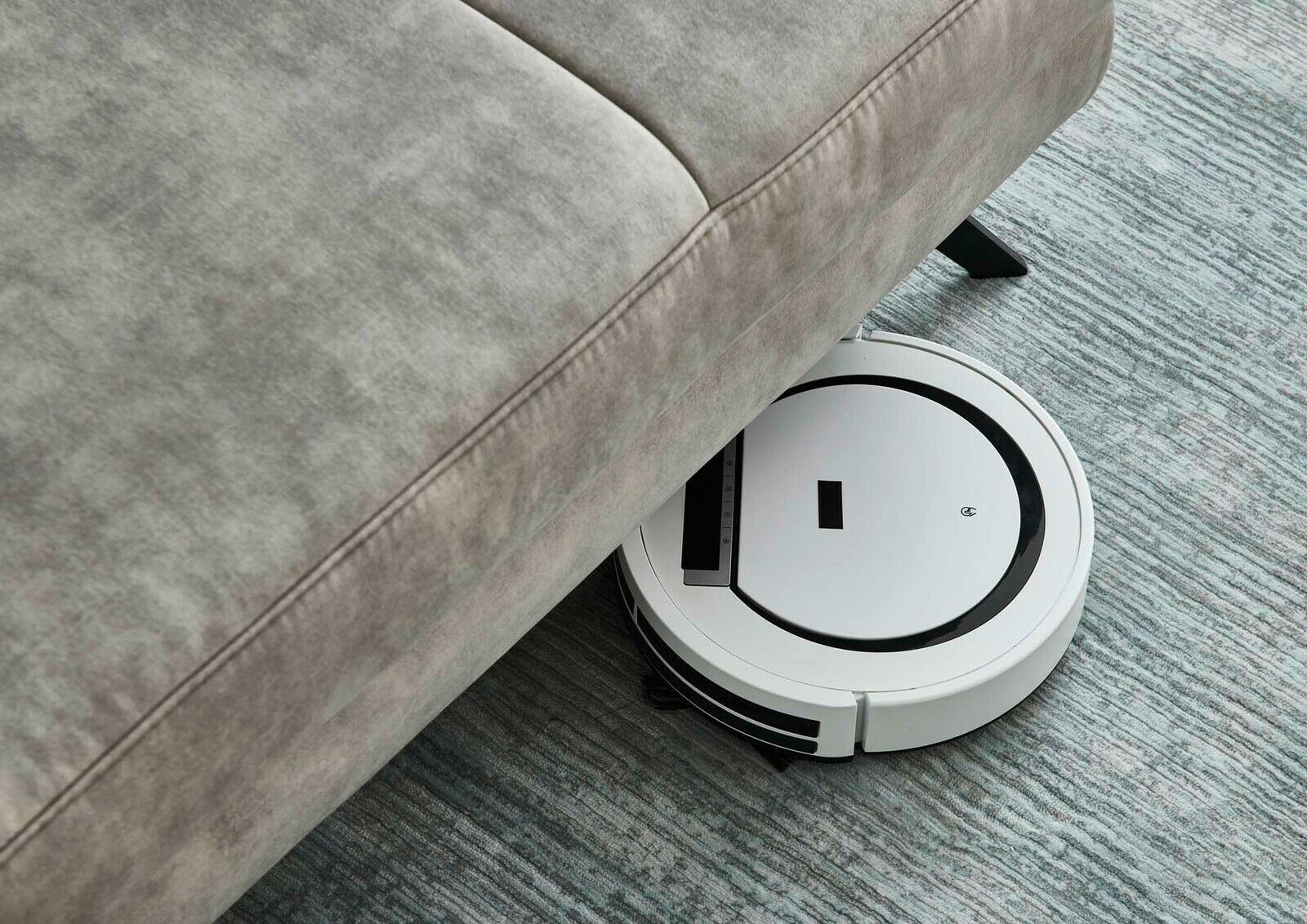Ein Saugroboter reinigt den Boden unter einer grauen Polsterrundecke. Perspektive von oben.