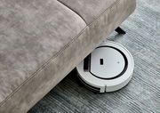Ein Saugroboter reinigt den Boden unter einer grauen Polsterrundecke. Perspektive von oben.