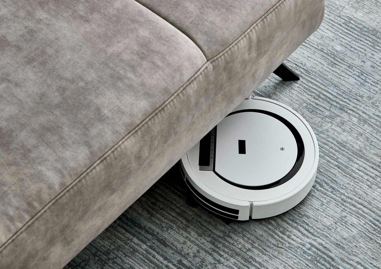 Ein Saugroboter reinigt den Boden unter einer grauen Polsterrundecke. Perspektive von oben.