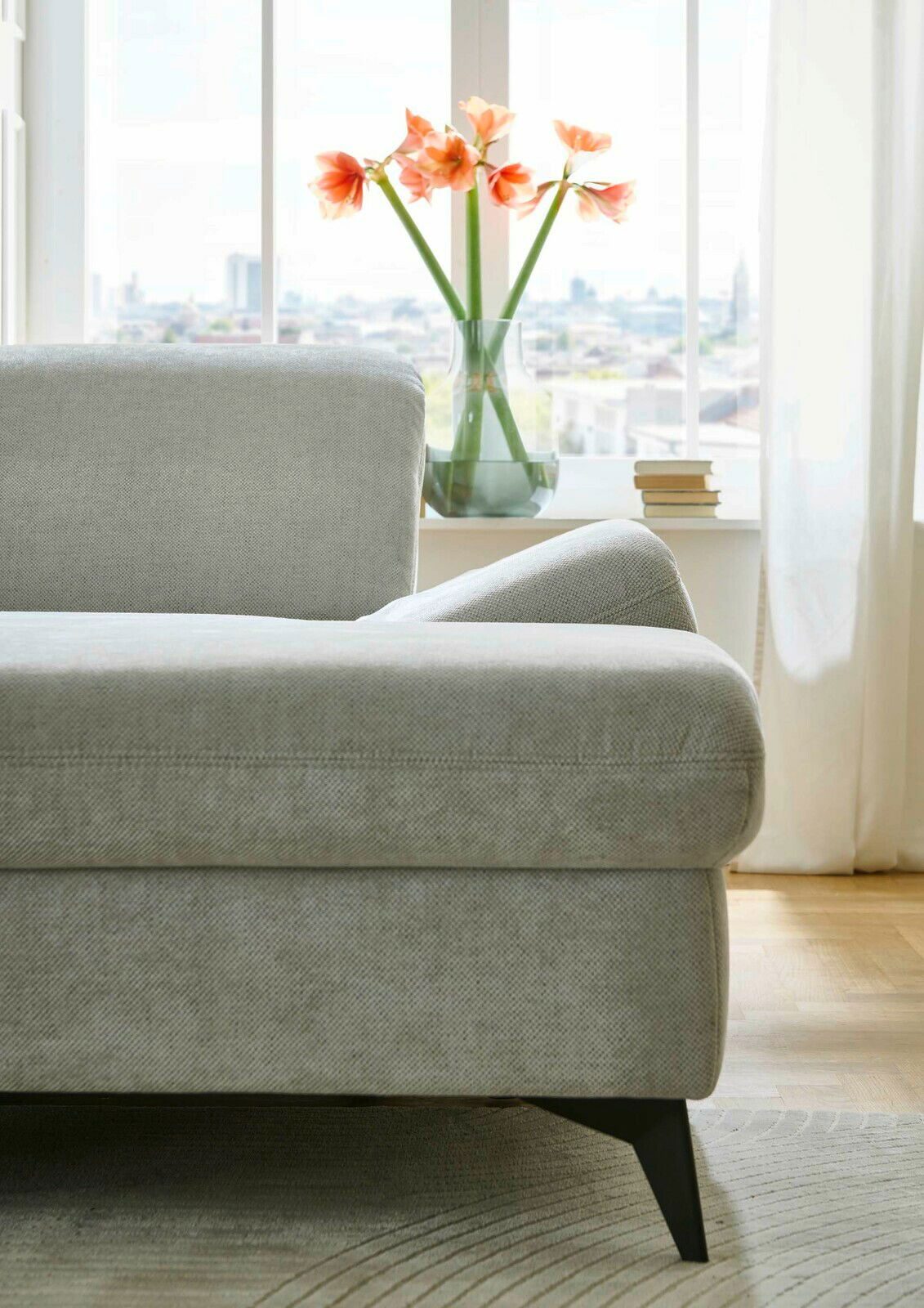 Graues Sofa in einer modernen Wohnung, seitliche Perspektive mit Blick auf ein Fenster mit Blumen und Büchern auf der Fensterbank.