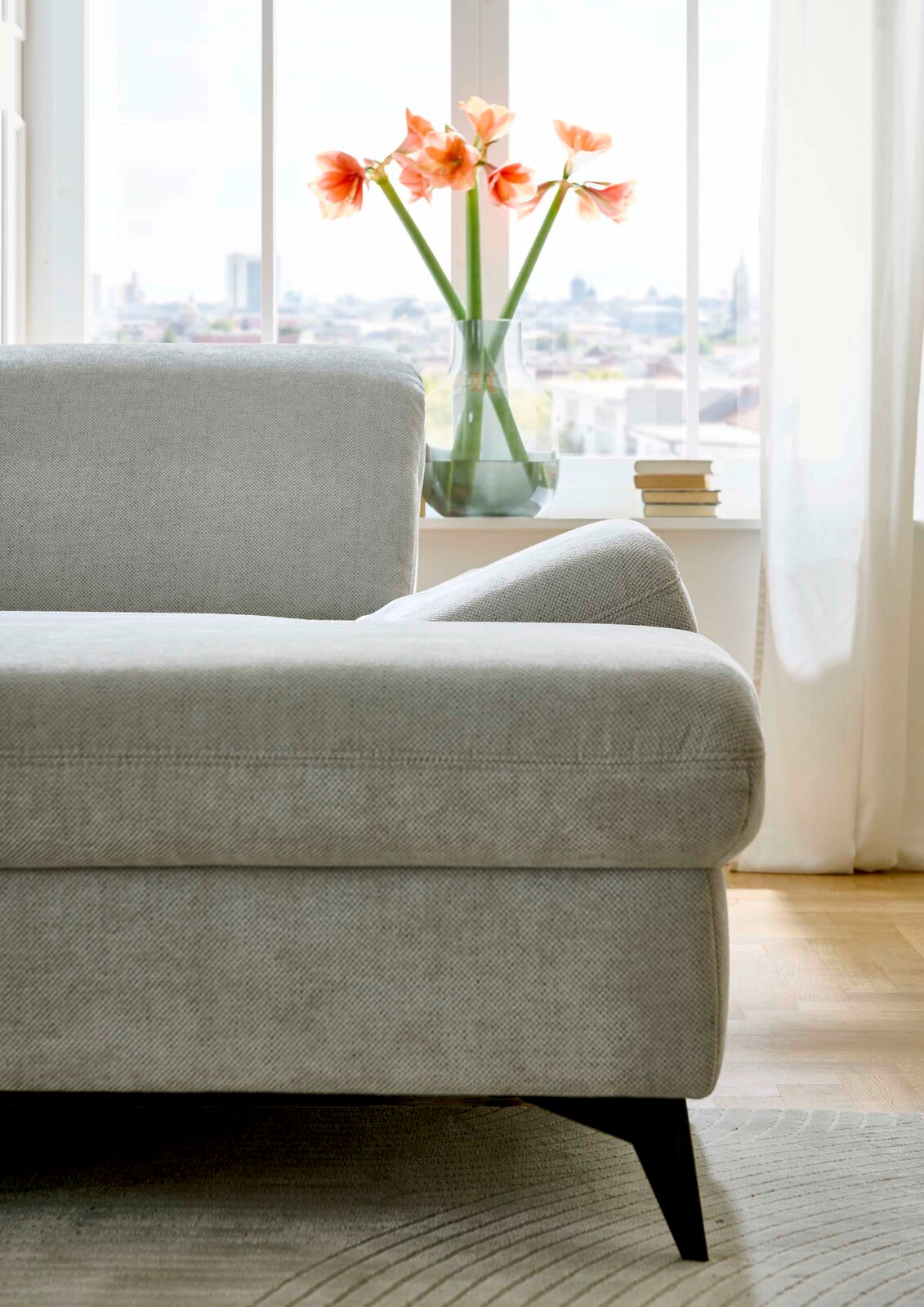 Graues Sofa in einer modernen Wohnung, seitliche Perspektive mit Blick auf ein Fenster mit Blumen und Büchern auf der Fensterbank.