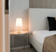 Moderne Tischleuchte mit weißem Lampenschirm auf einem ovalen Beistelltisch neben einem Bett, seitliche Perspektive.