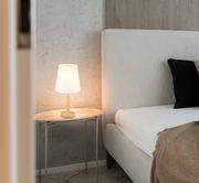 Moderne Tischleuchte mit weißem Lampenschirm auf einem ovalen Beistelltisch neben einem Bett, seitliche Perspektive.