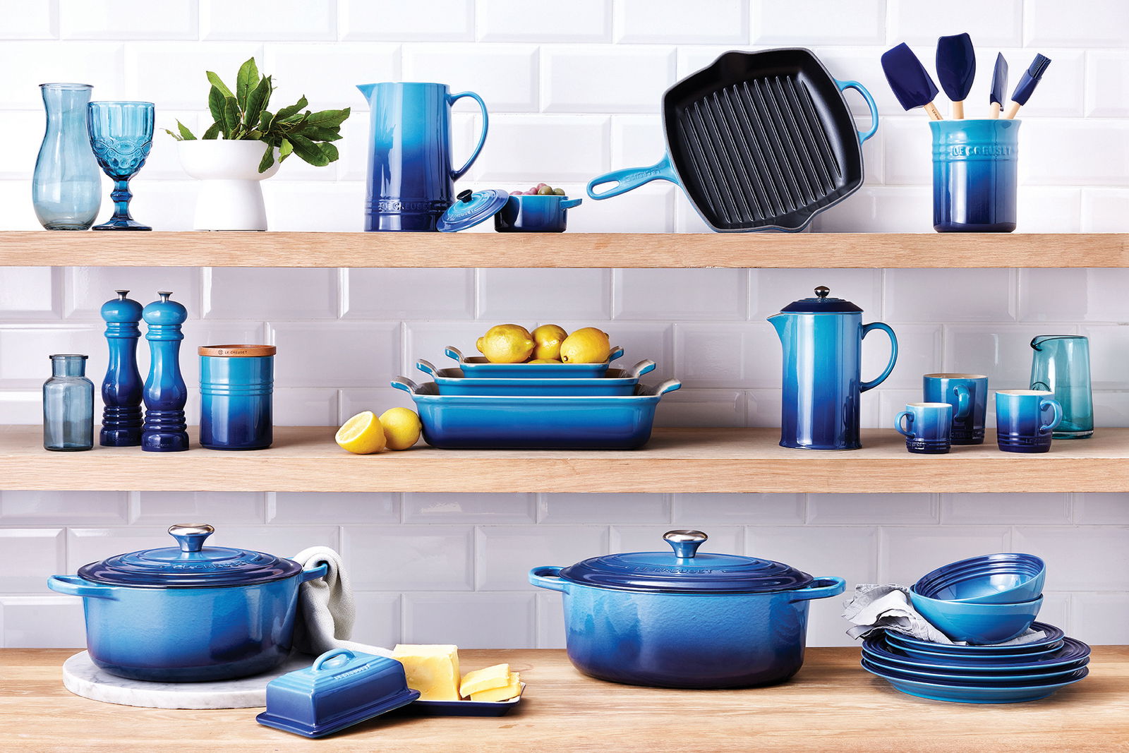 LE CREUSET Mini-Cocotte 4er-Set 10cm AZURE Verschiedene Küchenutensilien in Blau, darunter Mini Cocottes, auf Holzregalen vor einer weißen Fliesenwand, aus frontaler Perspektive.