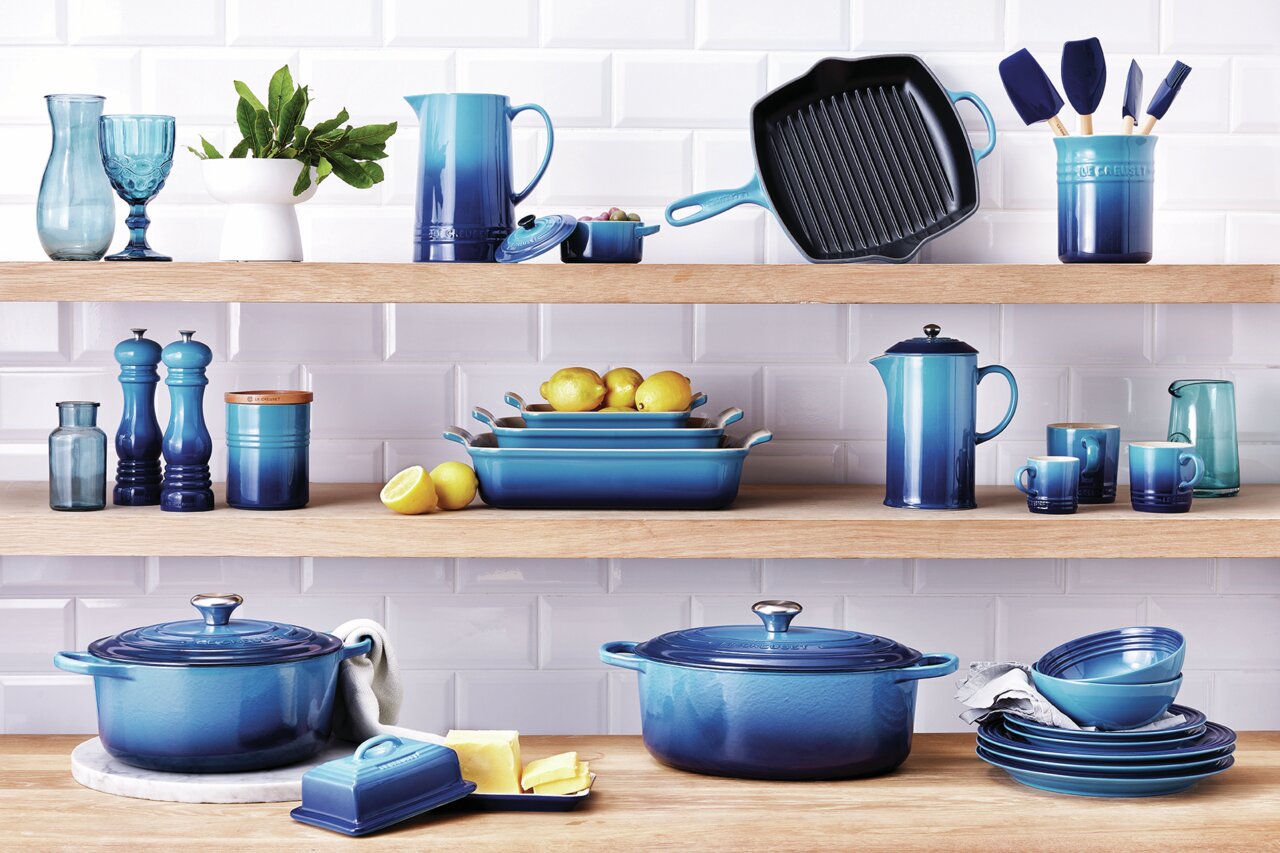LE CREUSET Mini-Cocotte 4er-Set 10cm AZURE Verschiedene Küchenutensilien in Blau, darunter Mini Cocottes, auf Holzregalen vor einer weißen Fliesenwand, aus frontaler Perspektive.