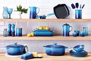 Verschiedene Küchenutensilien in Blau, darunter Mini Cocottes, auf Holzregalen vor einer weißen Fliesenwand, aus frontaler Perspektive.