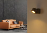 Moderne schwarze Wandleuchte mit zylindrischem Lampenkopf, warmem Lichtkegel nach unten, seitliche Wandmontage in Wohnraumszene; Perspektive: seitliche Ansicht leicht von vorn.