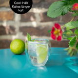 Doppelwandiges Thermo-Glas mit Limettenscheibe und Minzblatt, auf einem türkisfarbenen Tisch, vor einer Backsteinwand, aus seitlicher Perspektive.