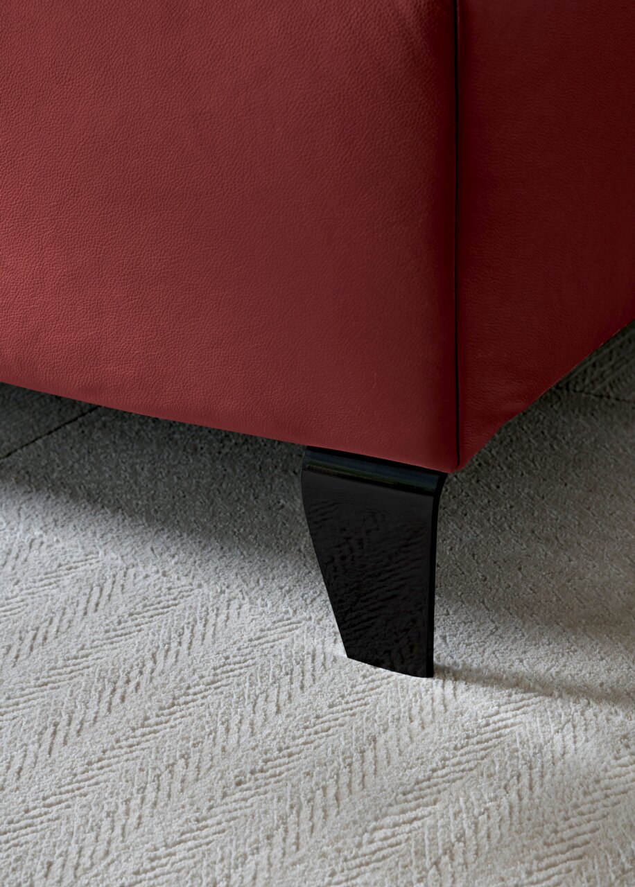 Nahaufnahme eines Boxspringbettbeins mit rotem Stoffbezug und schwarzem Holzfuß auf hellem Teppichboden, seitliche Perspektive