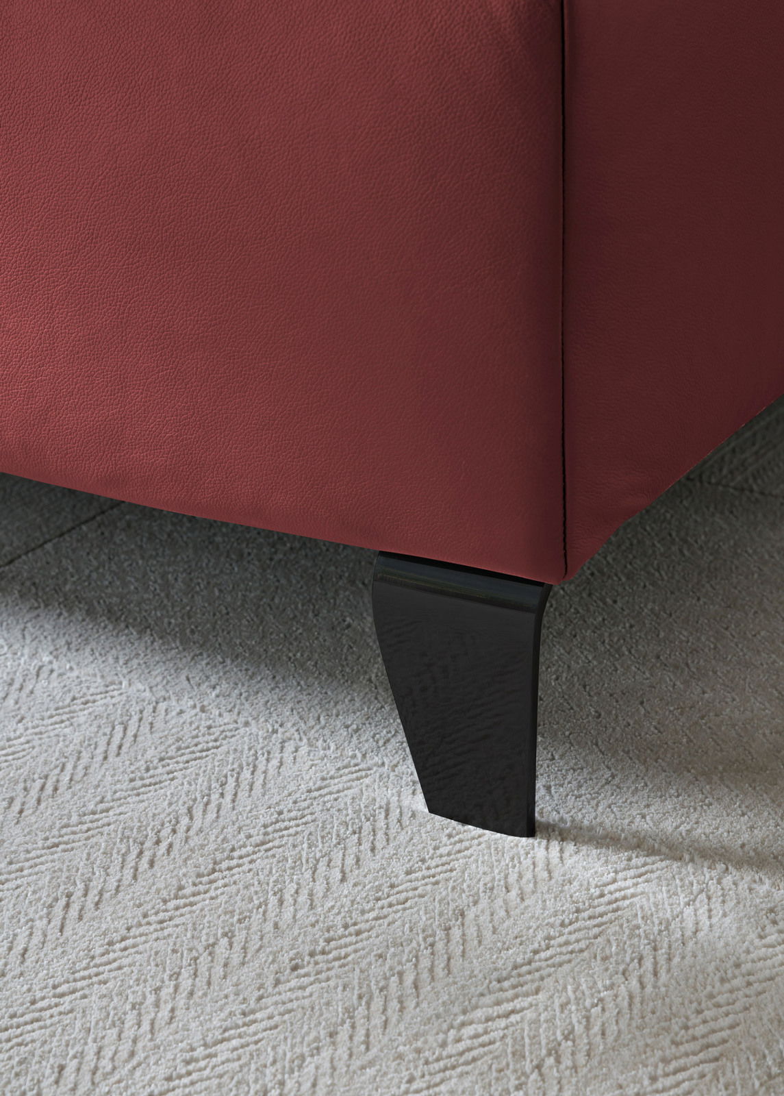 Nahaufnahme eines Boxspringbettbeins mit rotem Stoffbezug und schwarzem Holzfuß auf hellem Teppichboden, seitliche Perspektive