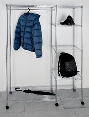 Silbernes, rollbares Kleiderregal aus Metall mit Seitenregal; blaue Steppjacke hängt am Bügel, oben schwarze Schuhe, unten schwarzer Rucksack und ein Schirm auf der unteren Ablage, Frontalansicht.