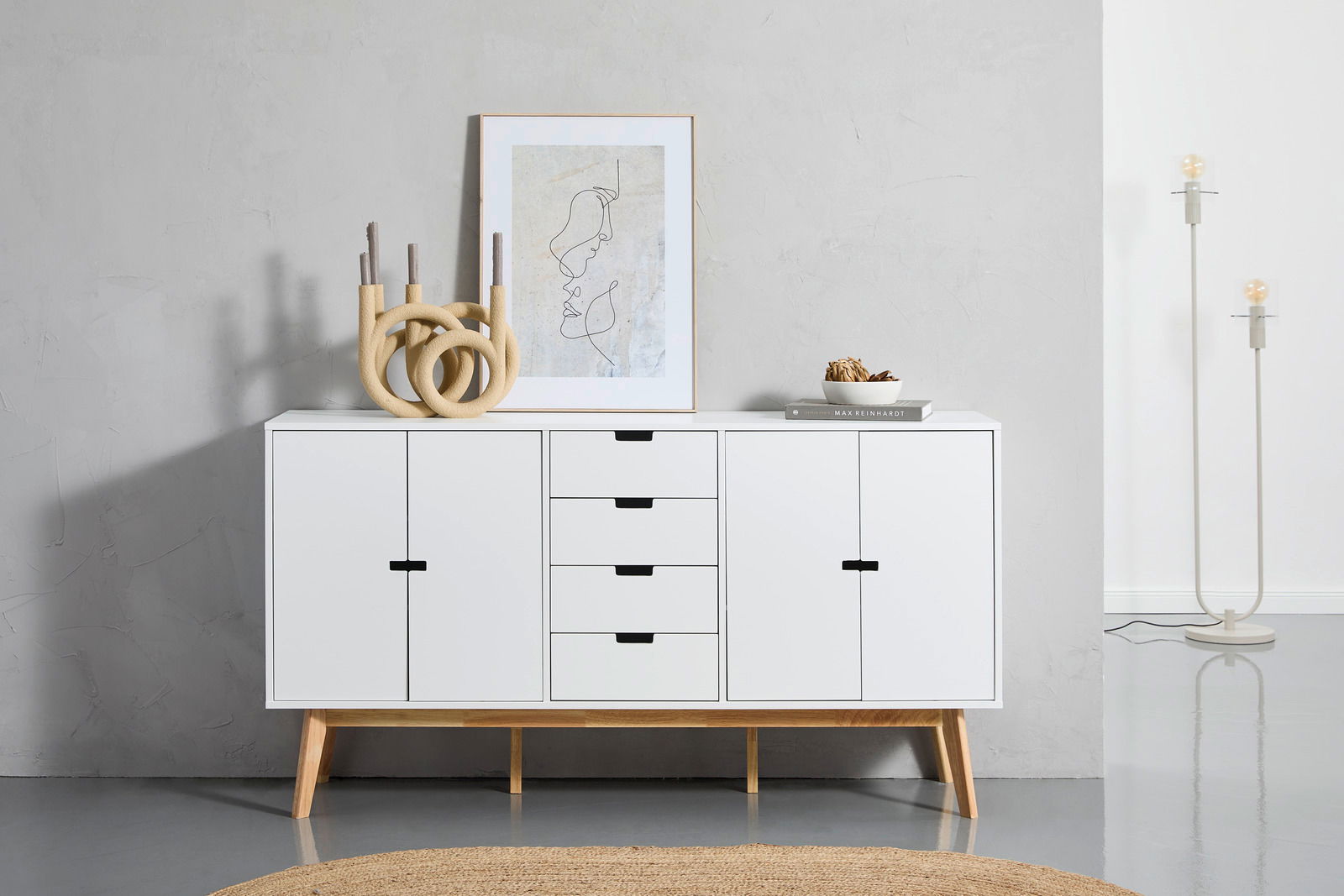 Weißes Sideboard mit Holzbeinen, frontal fotografiert, dekoriert mit Kunstwerk, Kerzenhalter und Schale.
