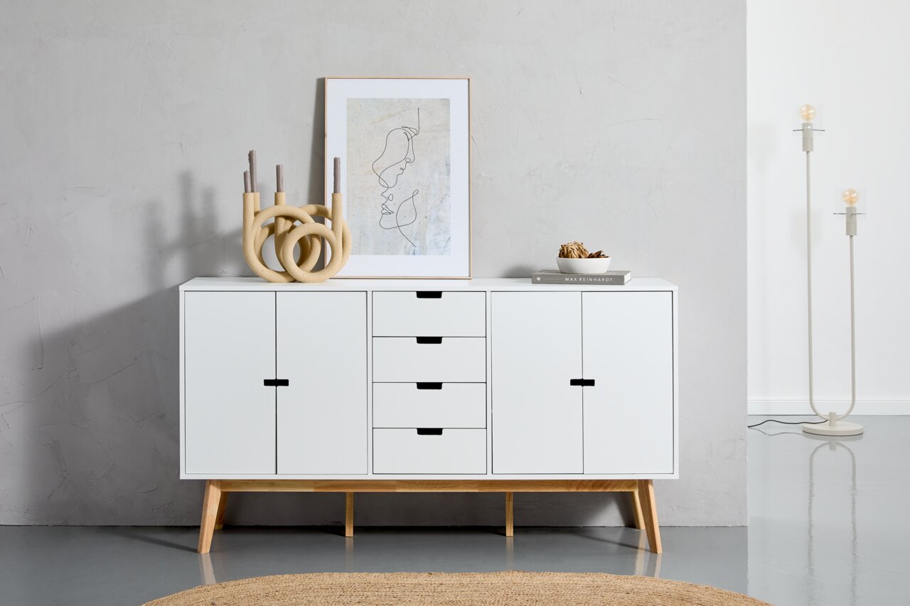 MID.YOU Dressoir LIPPON Wit dressoir met houten poten, gefotografeerd van voren, versierd met een kunstwerk, kandelaars en een schaal.