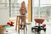 Kind in einem modernen Raum mit einem runden Holztisch und einem ergonomischen Sitzhocker für Kinder in Seitenansicht.