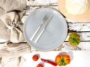Hellblauer Keramikteller mit braunem Rand, darauf Messer und Gabel; Draufsicht auf rustikalem Holztisch mit Stoffservietten, Tomaten, Chili und Teig auf Holzbrett.