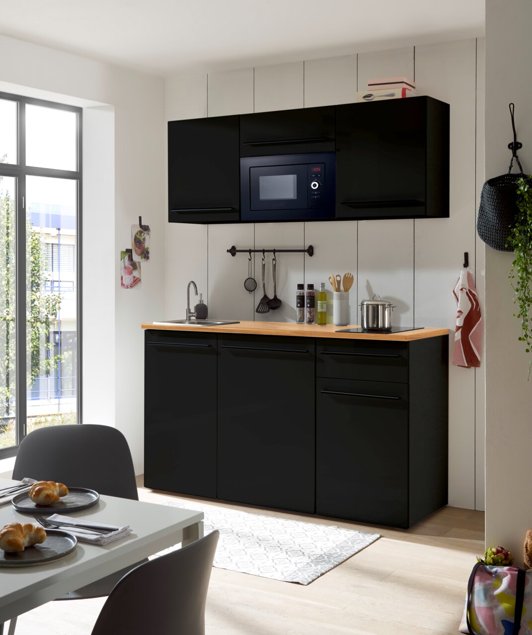 Moderne Single-Küche in schwarzem Design mit eingebauter Mikrowelle, Spüle und Arbeitsfläche aus Holz, frontal betrachtet.