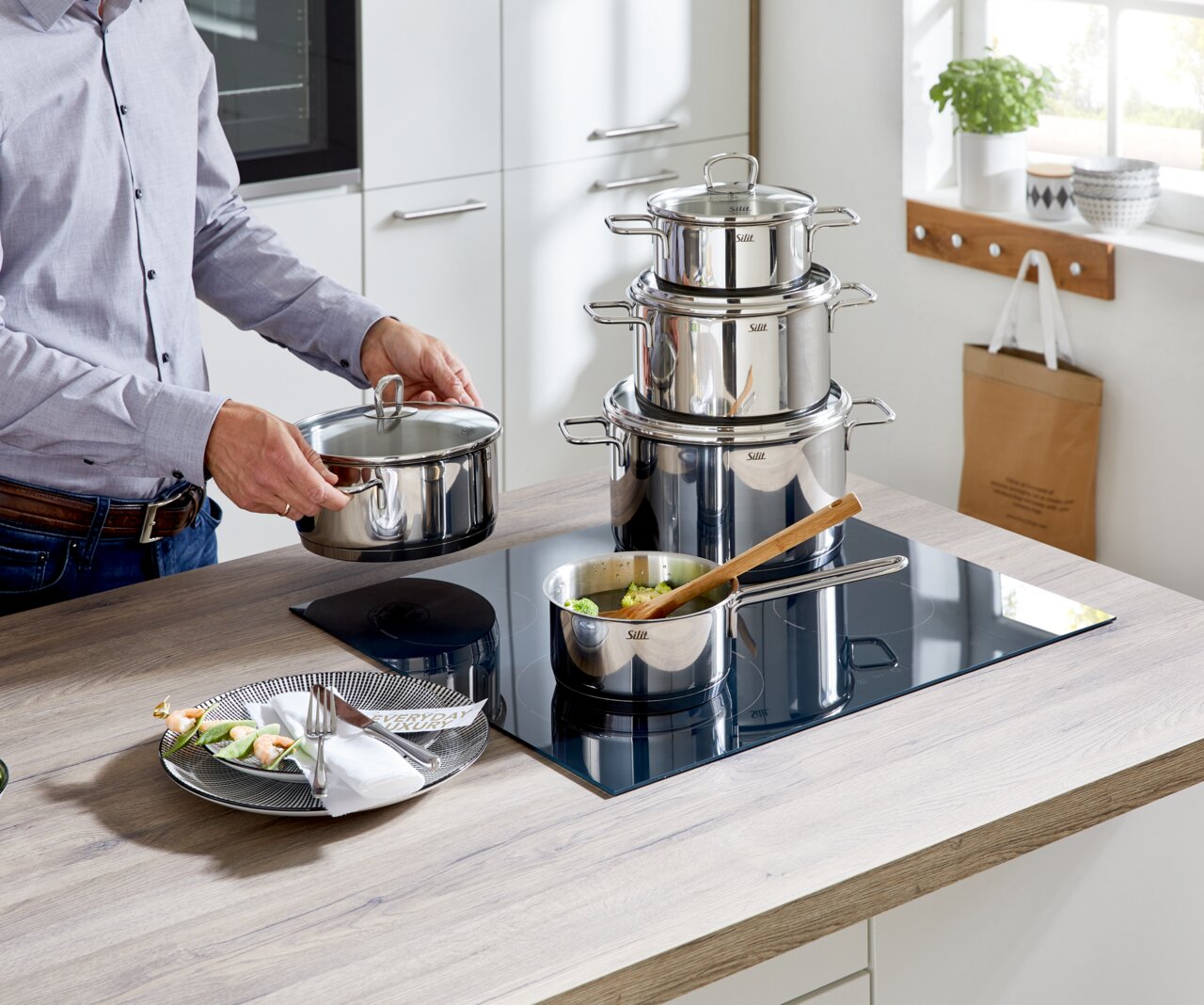 Küchenansicht mit einem 5-teiligen Topfset aus Edelstahl auf einem Herd, eine Person hält einen Topfdeckel, seitliche Perspektive.