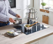 Küchenansicht mit einem 5-teiligen Topfset aus Edelstahl auf einem Herd, eine Person hält einen Topfdeckel, seitliche Perspektive.