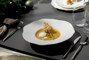 Weiße, modern geformte Suppenschale mit gelber Cremesuppe, Milchschaum und Spieß auf elegant gedecktem Tisch, schräg von oben fotografiert