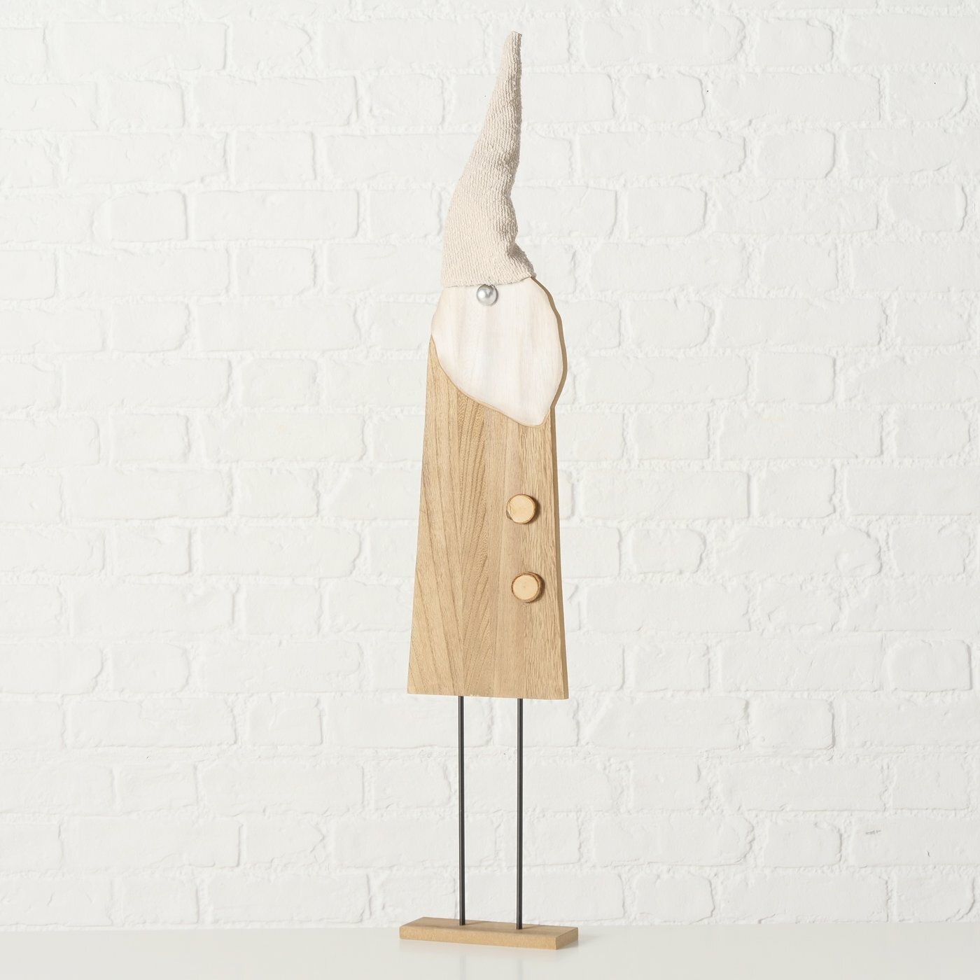 Holzfigur eines Gnomes mit spitzem Hut, frontal fotografiert vor einer weißen Ziegelwand.