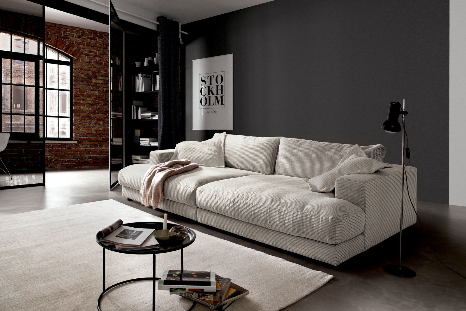 Pure Home Lifestyle Big Sofa PURE 979 Modernes, cremefarbenes Megasofa in einem stilvollen Wohnzimmer mit Ziegelwand und großen Fenstern, seitliche Perspektive.