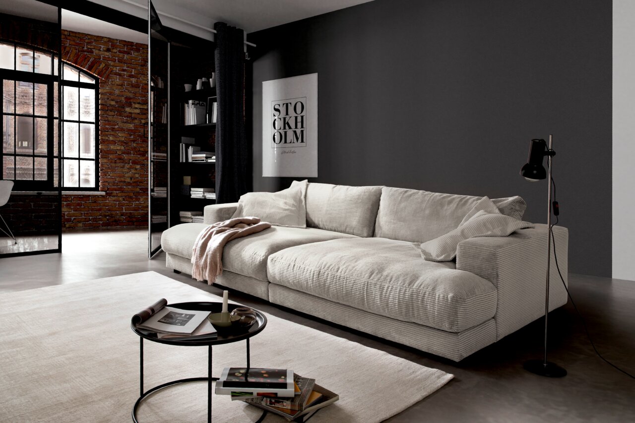 Modernes, cremefarbenes Megasofa in einem stilvollen Wohnzimmer mit Ziegelwand und großen Fenstern, seitliche Perspektive.