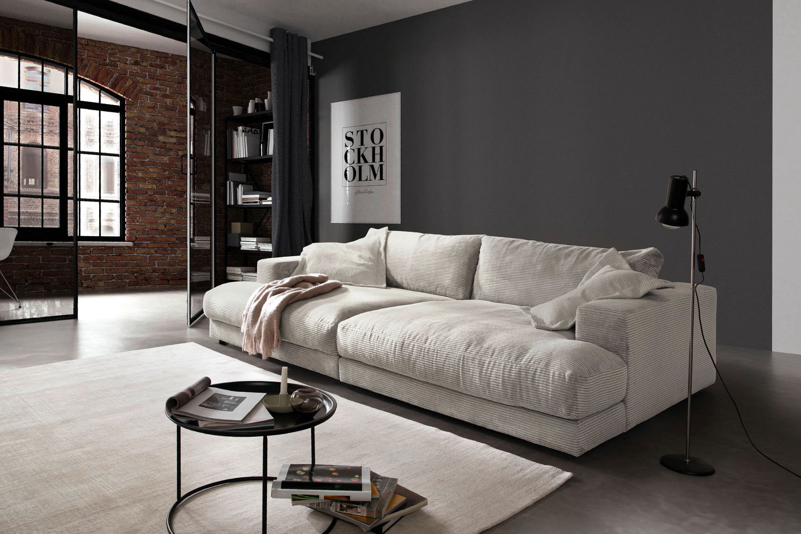 Modernes, cremefarbenes Megasofa in einem stilvollen Wohnzimmer mit Ziegelwand und großen Fenstern, seitliche Perspektive.