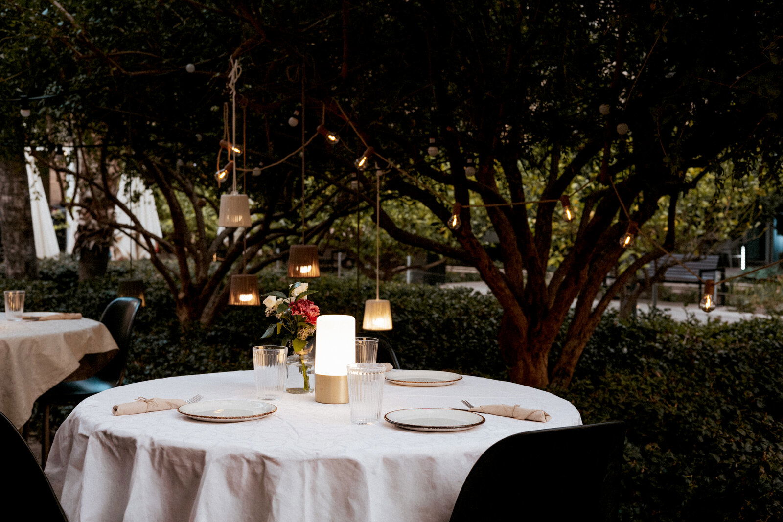 Rund gedeckter Outdoor-Esstisch mit weißer Tischdecke, Teller und Gläsern, zentraler LED-Tischlampe und Blumen, umgeben von Bäumen mit Lichterketten, aufgenommen aus leicht erhöhter Frontperspektive.