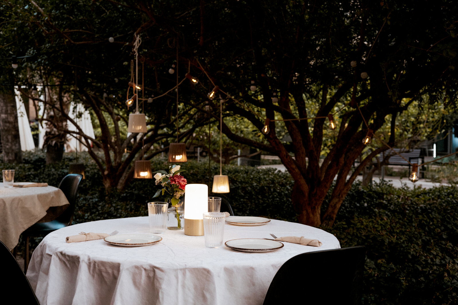 Rund gedeckter Outdoor-Esstisch mit weißer Tischdecke, Teller und Gläsern, zentraler LED-Tischlampe und Blumen, umgeben von Bäumen mit Lichterketten, aufgenommen aus leicht erhöhter Frontperspektive.