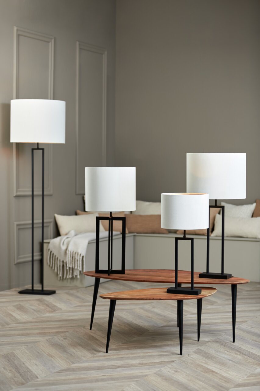 Vier moderne Lampen mit weißen zylindrischen Lampenschirmen und schwarzen Ständern, in einem stilvollen Raum mit Holzboden und neutralen Wänden, aus einer seitlichen Perspektive fotografiert.