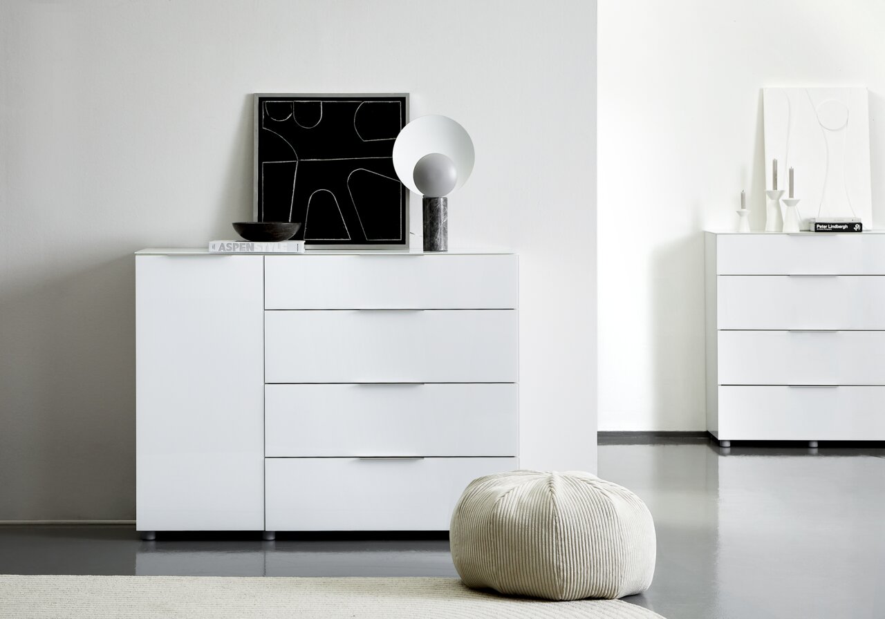 Weiße Kombikommode mit vier Schubladen und einer Tür, dekoriert mit einem modernen Kunstwerk und einer Lampe, aus der Frontalperspektive fotografiert.
