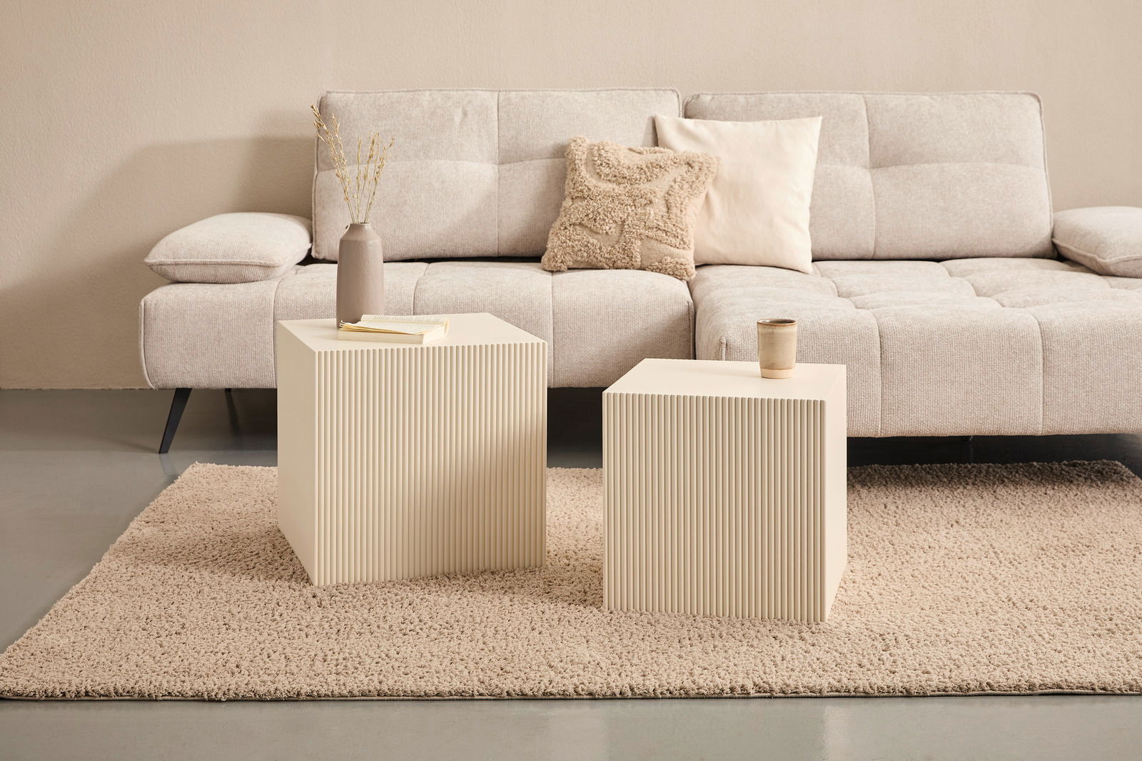 Zwei cremefarbene Beistelltische mit geriffelter Oberfläche vor einem beigen Sofa, seitliche Perspektive.