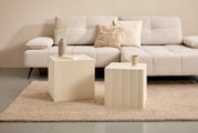 Zwei cremefarbene Beistelltische mit geriffelter Oberfläche vor einem beigen Sofa, seitliche Perspektive.
