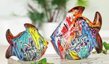 Zwei bunte Fischskulpturen im Street-Art-Stil auf einem Tisch, seitliche Perspektive
