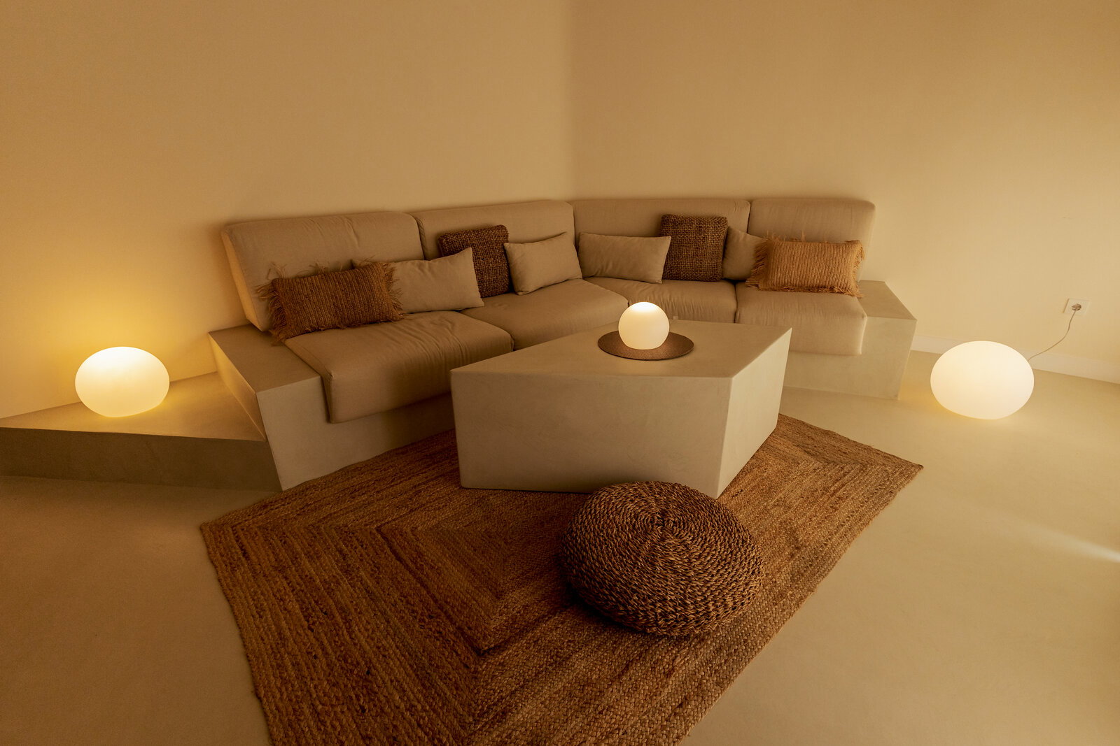 Gemütliche, minimalistische Wohnzimmerecke mit beigem Ecksofa und Kissen, kubischem Couchtisch, Jute-Teppich und runder Bodenleuchte in warmem Licht, aufgenommen aus leicht erhöhter Frontperspektive.