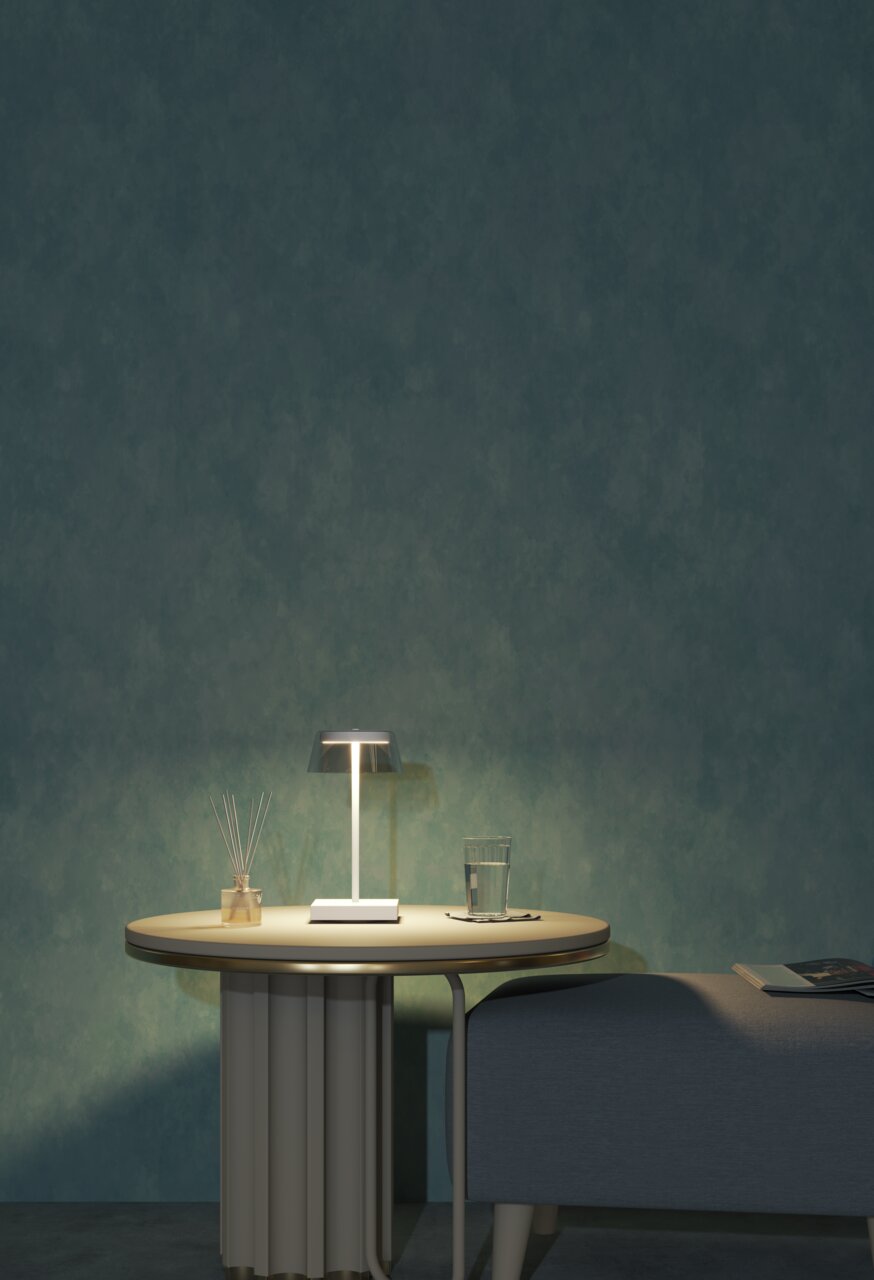 Moderne Akku-Tischleuchte auf einem runden Tisch, seitliche Perspektive, mit einem Glas Wasser und einem Duftstäbchen-Set daneben.