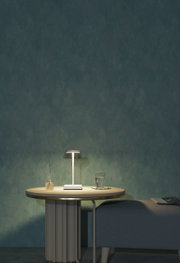 Moderne Akku-Tischleuchte auf einem runden Tisch, seitliche Perspektive, mit einem Glas Wasser und einem Duftstäbchen-Set daneben.