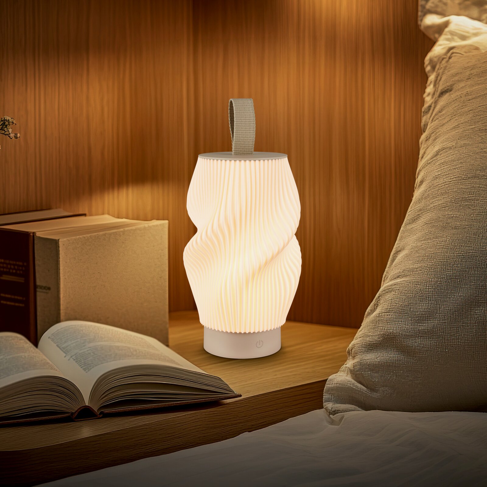 Leuchtende, tragbare LED-Tischlampe in weiß mit geriffeltem, gedrehtem Schirm und Trageschlaufe auf einem Nachttisch neben Buch und Bett, Frontansicht auf Augenhöhe