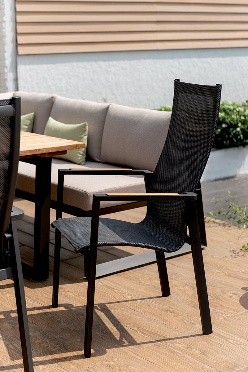 Moderne zwarte stapelstoel met hoge rugleuning en houten armleuningen, gezien vanaf de zijkant op een terras naast een houten tafel en een bank.